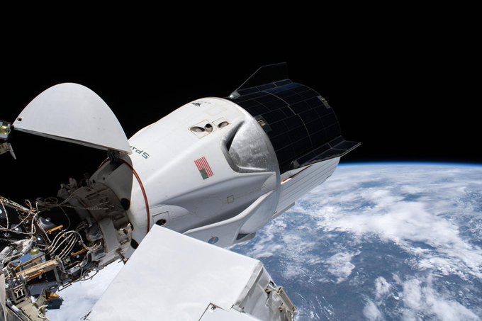 Crew Dragon docked to the space station. It juts out fromt he docking port and Earth is below it. The darkness of space is the background.