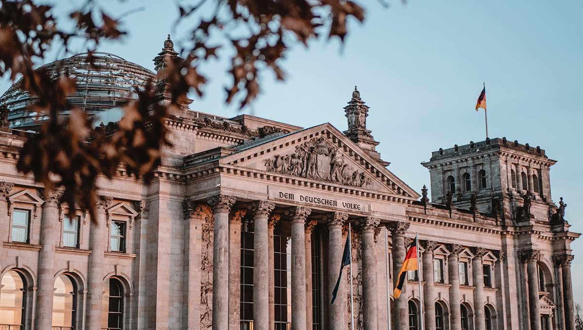 Auf dem Bild ist das Reichstagsgebäude abgebildet. Im Vordergrund ist unscharfes Laub zu sehen. 