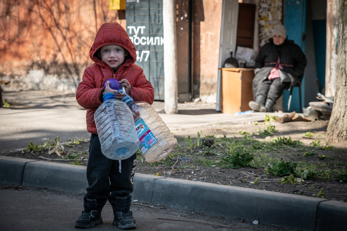 A young boy clutches his ball and water bottles outside his destroyed apartment in #Mariupol