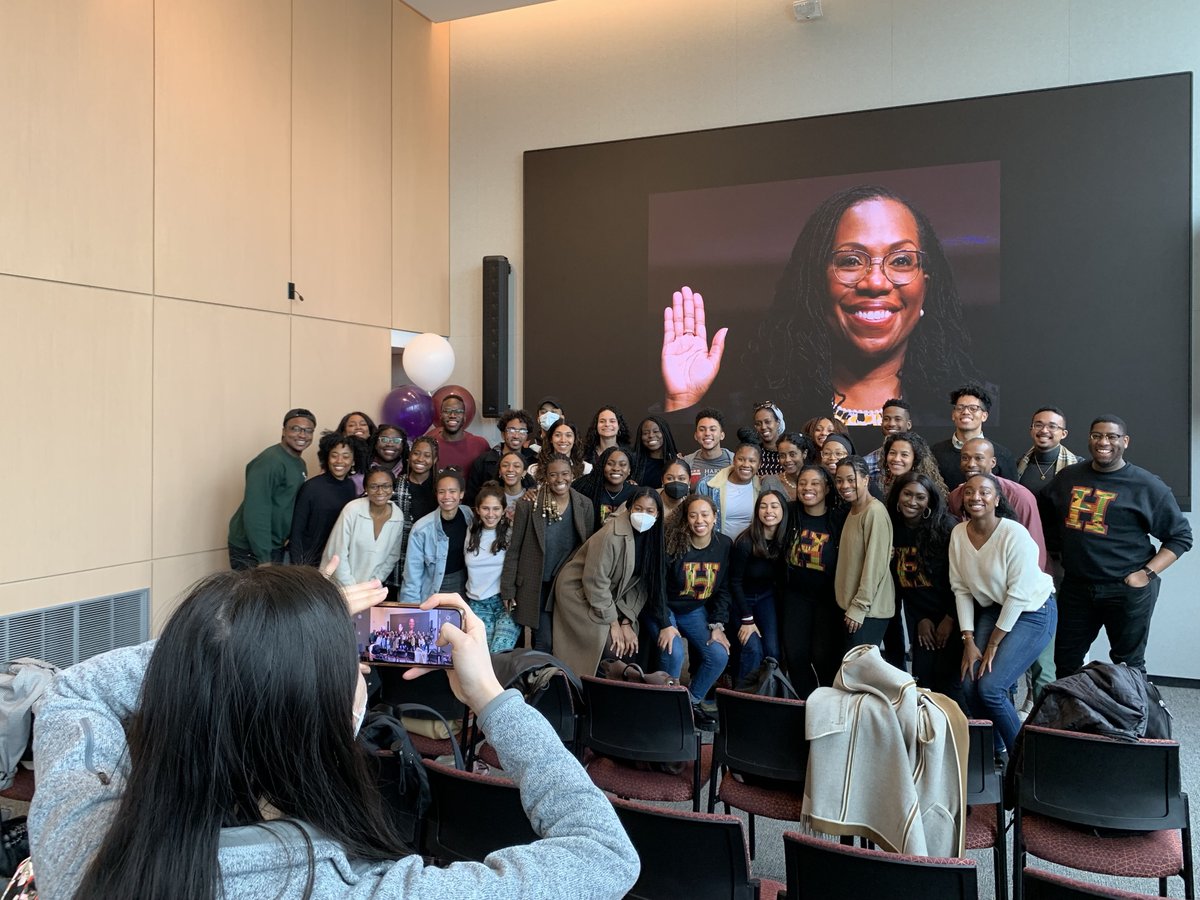 A group of students pose in front of a large screen with an image of Ketanji Brown Jackson. Another student is taking a photo of them with a mobile phone.