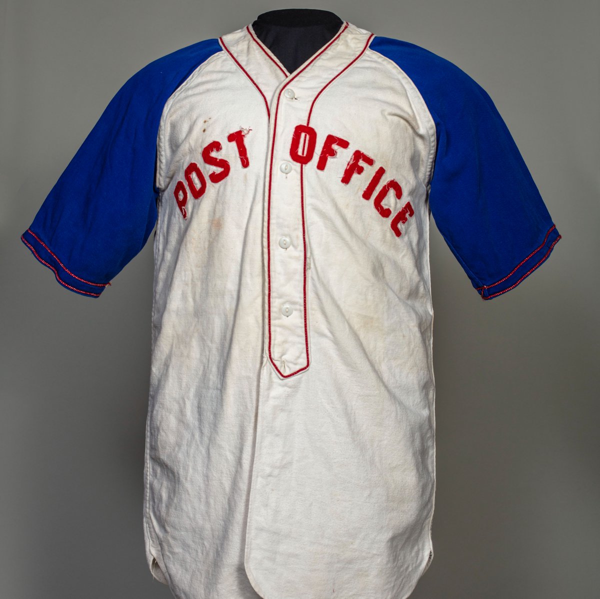 Baseball jersey with white body, blue arms, and red piping around chest buttons and neck. Red text: Post Office. 