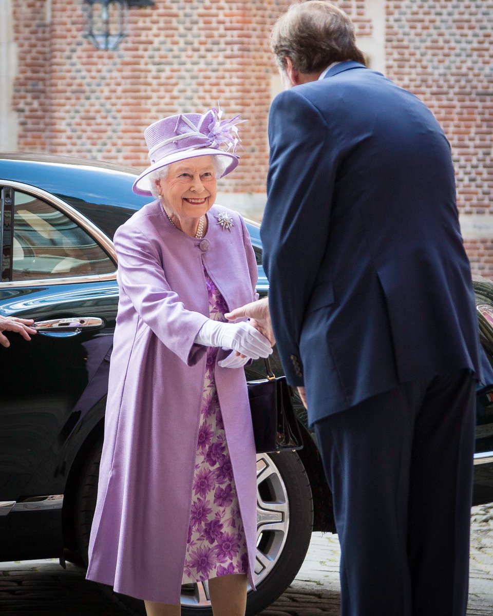 Her Majesty The Queen wearing purple shaking hands with a gentleman with his back to the camera.
