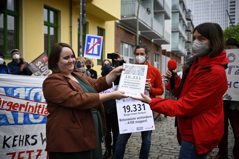 Eine Mitarbeiterin von Campact, die eine rote Jacke und einen Mund-Nasen-Schutz trägt, übergibt eine Petition mit 19.331 Unterzeichnungen gegen den Bau der A100 in Berlin an Ricarda Lang, eine der Bundesvorsitzenden von Bündnis 90/Die Grünen. Ricarda Lang trägt einen braunen Blazer und lächelt freundlich. Im Hintergrund stehen Demonstrant*innen mit Bannern und Schildern gegen den Bau der Autobahn. 