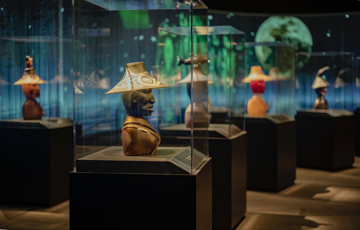 Museum gallery with five cases, dramatically lit with green and blue background. Bust-length figured in each case wear wide hats.