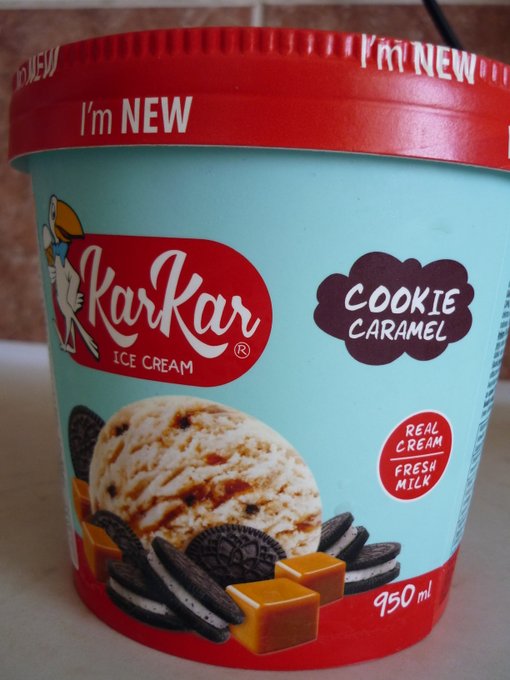 Kar Kar Cookie Caramel ice cream seen from the side. The lid has two 'I'm NEW' strips on the side. The 'bucket' shows funny logo with a cartoonish bird holding an ice cream cone, a 'presentation' suggestion image showing a scoop surrounded by caramels and oreo knockoffs (way, way nicer nicer than actual oreos), ice cream type (cookie caramel), a sticker like button that reads 'real cream, fresh milk' and quantity (950ml).