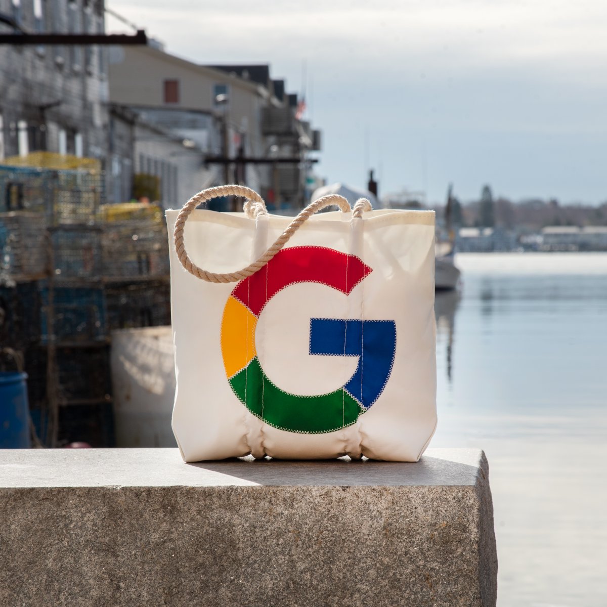 A tote bag is perched on a ledge in front of a harbor. The Google Super G is stitched on to the front of the bag.