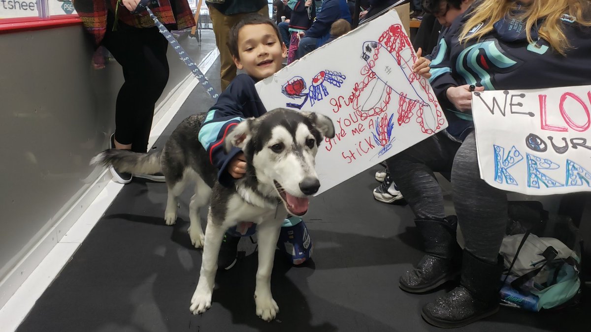 Child holding sign posing with husky mix dog. Hockey fans in the background.