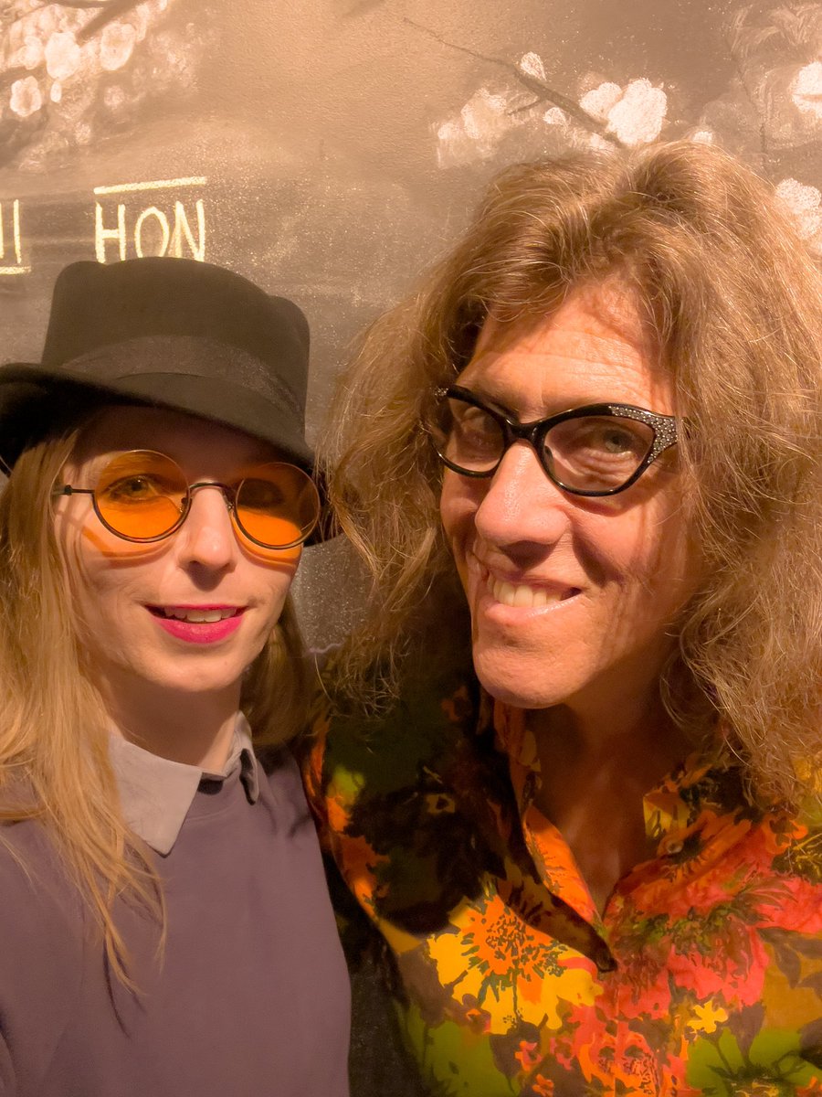 Chelsea E. Manning to the left wearing orange sunglasses and a cat ear bowler hat, with Susan Stryker wearing cat eye glasses and a floral blouse.