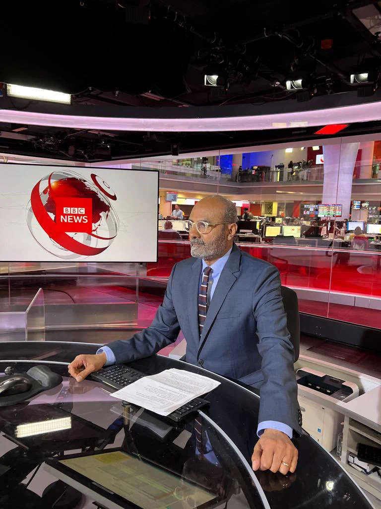 George Alagiah looks in to the camera. He sits behind the BBC News desk in the studio.