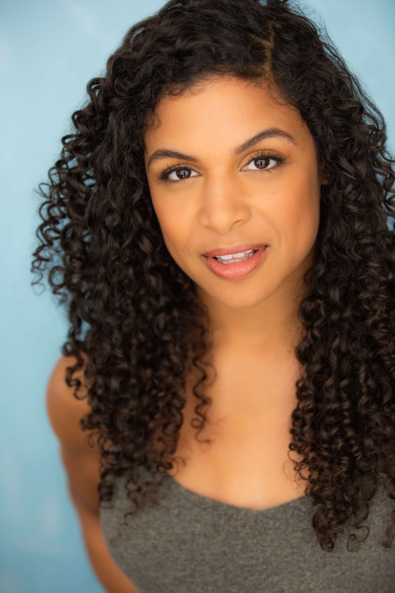 A headshot of actress and choreographer Kristin Yancy. She stands in front of a light blue background wearing a gray tank top. She has long, curly brown hair going past her shoulders.