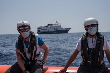 2 Personen auf dem Rand eines RHIBs auf dem Meer. Zwischen ihnen hindurch ist im Hintergrund die GEO BARENTS zu sehen, das aktuelle MSF-Rettungsschiff im zentralen Med.