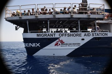 Das Heck der PHOENIX von MOAS;<br>auf dem Deck und einer darüber liegenden Plattform drängen sich Menschen.<br>Auf der Reling steht 'Migrant Offshore Aid Station',<br>darunter das Logo von MSF.<br>[MSF stellte 2x in 2015 das medizinische Personal für Mission der PHOENIX]