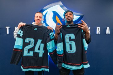 macklemore & marshawn lynch holding two blue kraken jerseys that they signed for each other