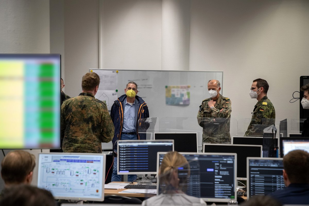 Generalmajor Jürgen Setzer steht mittig, links neben ihm ist Herr MdB Müller, rechts neben ihm ein Soldat. Sie hören einem/r SoldatIn, der/die vor ihnen steht zu. Im Vordergrund sind PCs und Menschen an PCs sitzend leicht verschwommen wahrzunehmen. Alles befindet sich in einem großen Raum. 
