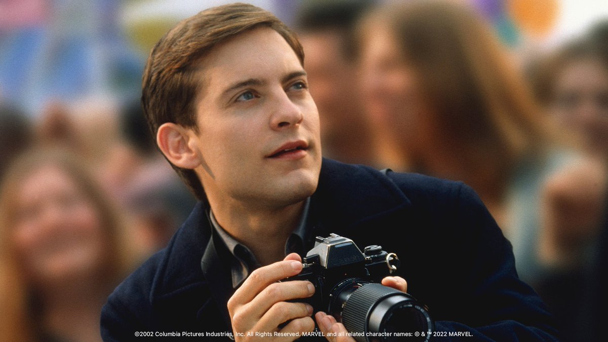 In this still, Peter Parker (portrayed by Tobey Maguire) stands with his professional camera poised beneath his chin, ready to be lifted up to his eyes at a moment’s notice. He’s wearing a blue coat and a grin on his face.
