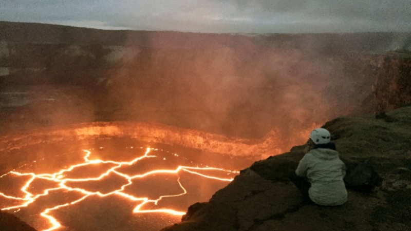 A person in a hooded jacket and hat sits on the edge of an active volcano which has lava inside it.