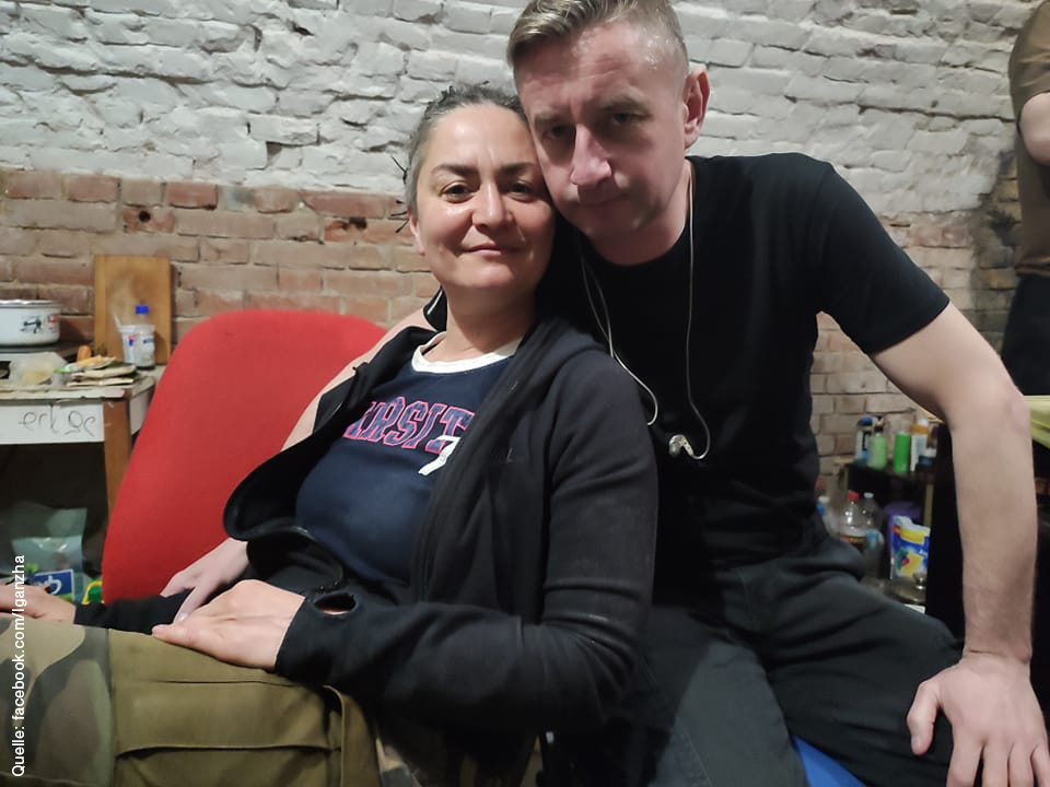 Lesia Gandza and Serhij Zhadan sitting next to each other in bomb shelter in Charkiw. She is leaning against him. She wears military trousers and a black jacket. He wears a black t-shirt. Headphones hanging around his neck. In the background is a improvised cooking zone and some bottles.