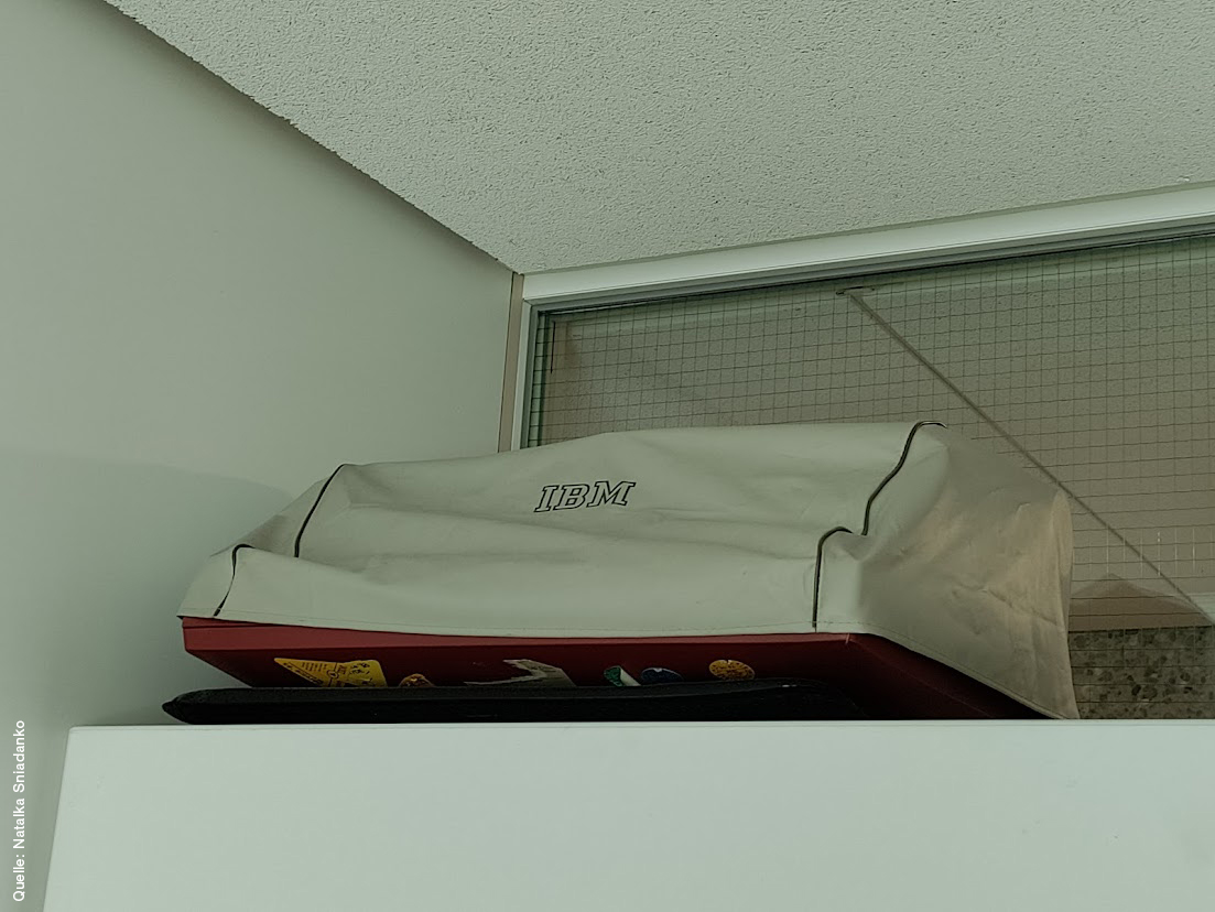 A typewriter stands on top of a cabinet. It’s covered with a dust cover. On the cover is written IBM.