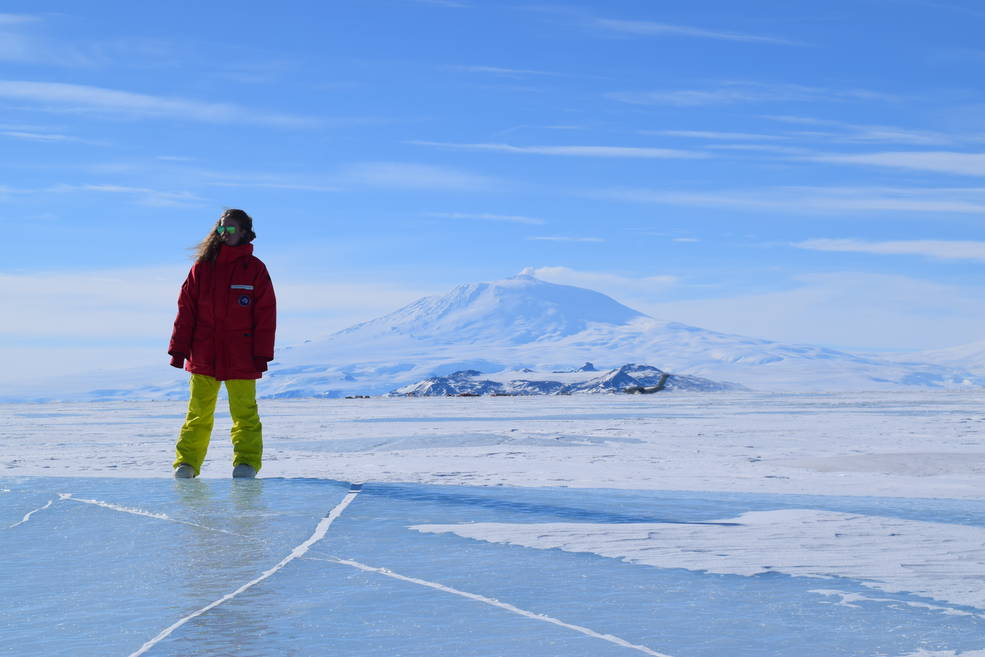 Woman in a red parka standing on top of ice in Antarctica. She is off-center, and the view is mostly of the ice and snow in Antarctica. In the background is a snow-covered hill and the sky is blue, indiciating it is dry and freezing. 