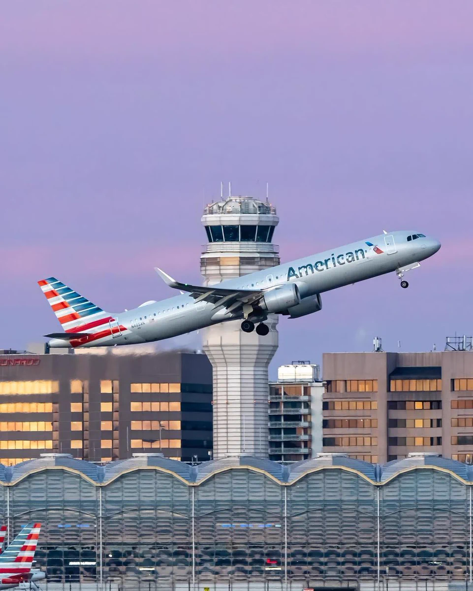 American Airlines plane taking off from Reagan National Airport. Tower and Terminal are visible in the background. The sky is purple and it's just after sunset. 