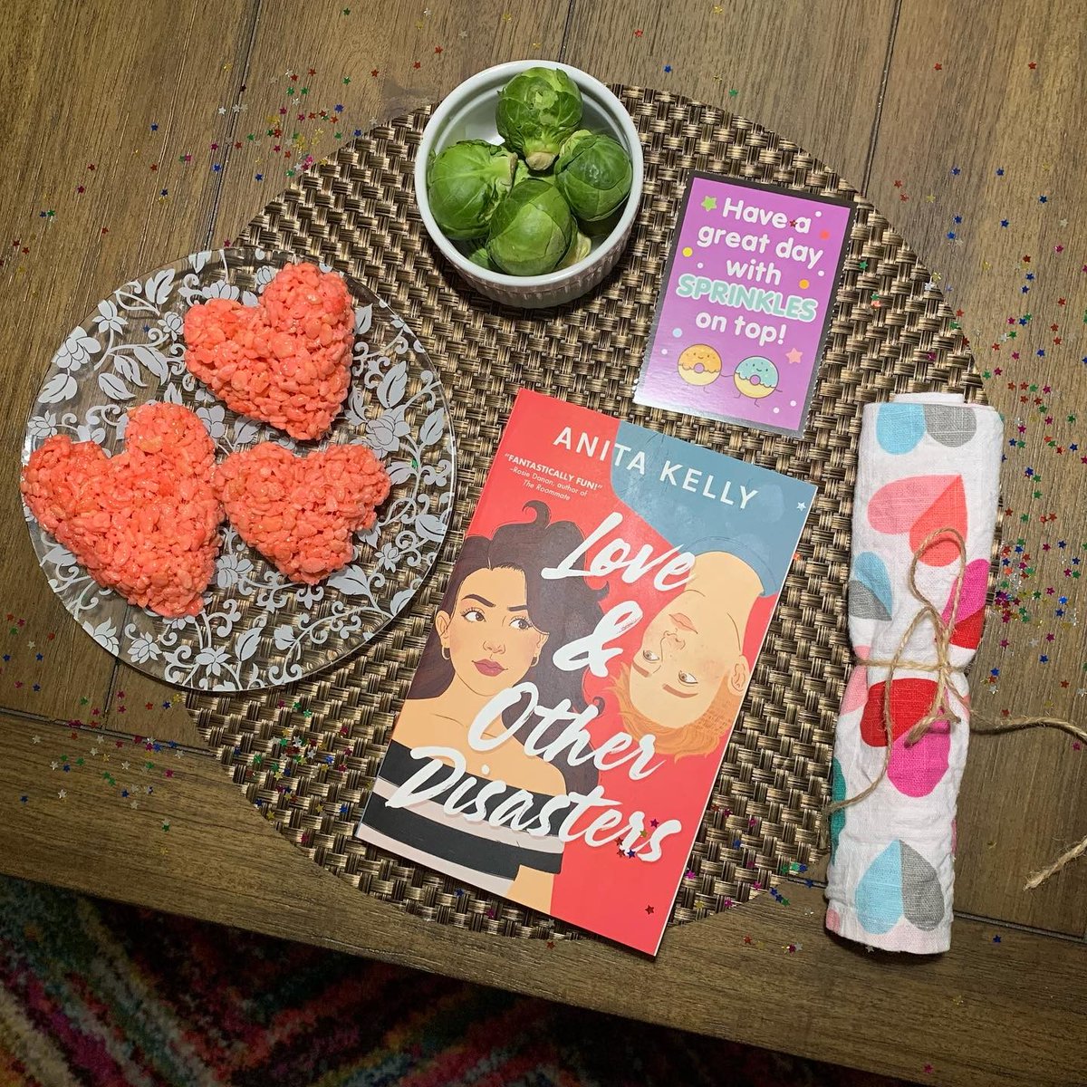 Love and Other Disasters by Anita Kelley on a table next to heart shaped rice-krispy treats, a bowl of brussels sprouts and a note that says "Have a great day with sprinkles on top."