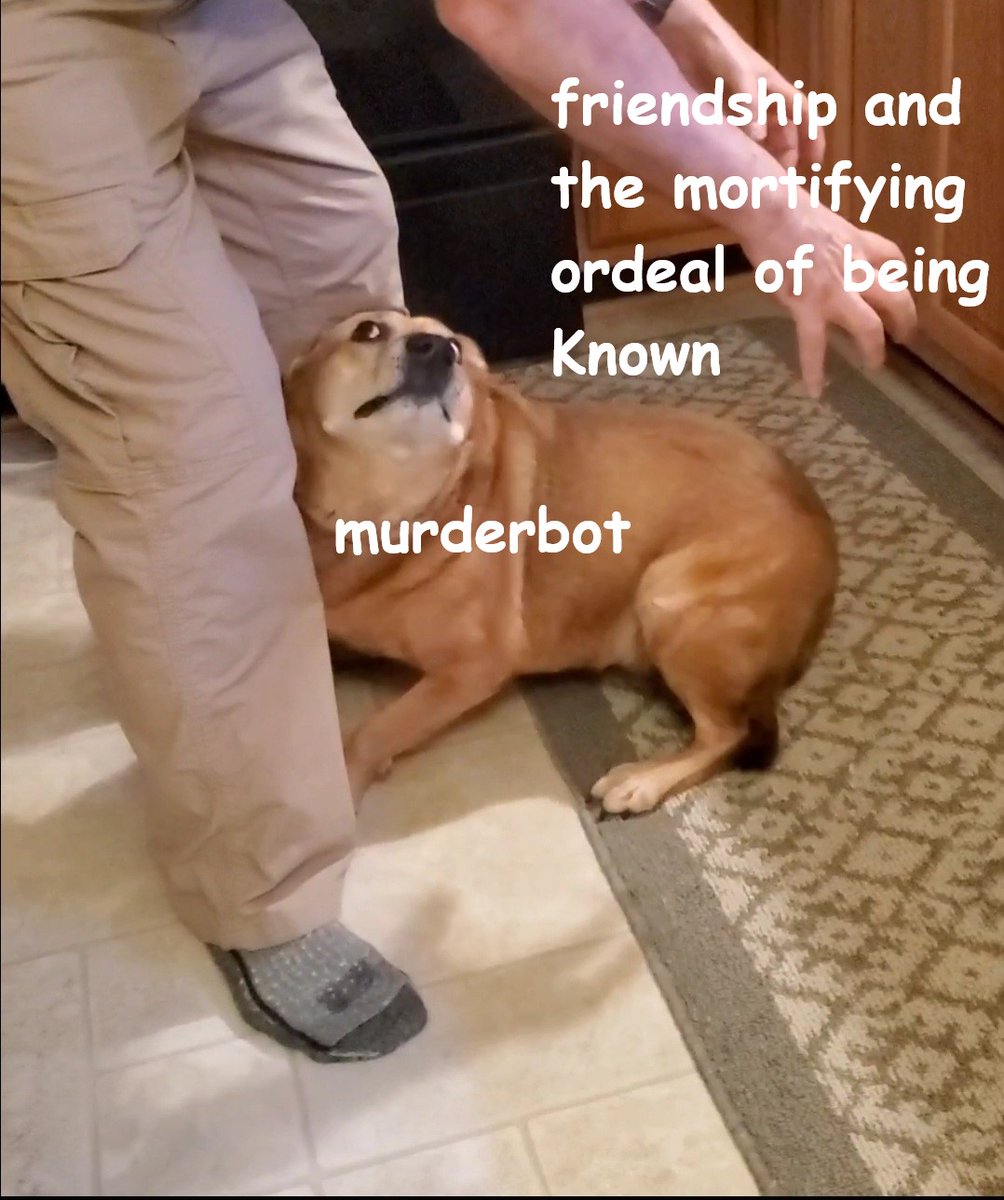 a reddish-brownish dog low to the ground as a person wearing khaki pants stands over her. Only the persons legs and arms can be seen as he's reaching down to grab the dog's butt. The dog is staring at the person with the whites if her eyes showing and her black lips kind of making a < sort of shape. the hands labeled 'friendship and the mortifying ordeal of being known' and the dog labeled 'murderbot'