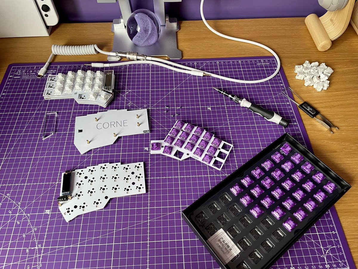 Dismantled components on purple desk mat. Box of purple switches, loose white switches, screwdriver, switch puller, case base, plate, pcb, and oled guard.