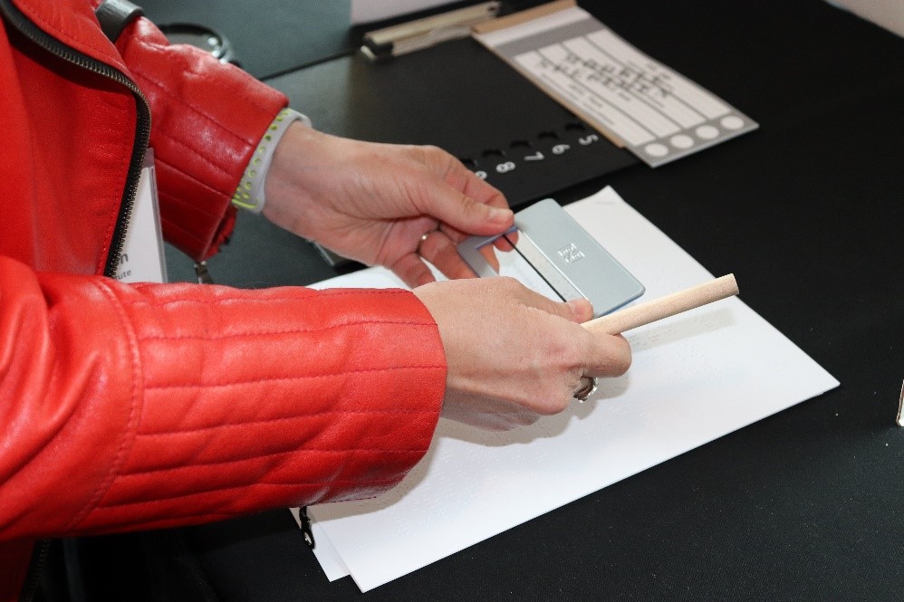 [Image description: A woman holds a signature guide and a pencil over a white page. A tactile ballot overlay is next to her.] 