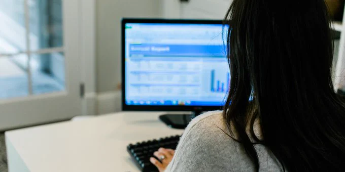 Over-the-shoulder view of a person sitting at a home office desk, typing on a keyboard, and looking at a large computer monitor
