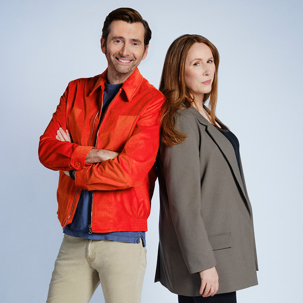 David Tennant and Catherine Tate stand back to back as they return to Doctor Who. David Tennant wears a red jacket over a blue t-shirt and Catherine Tate wears a grey blazer.