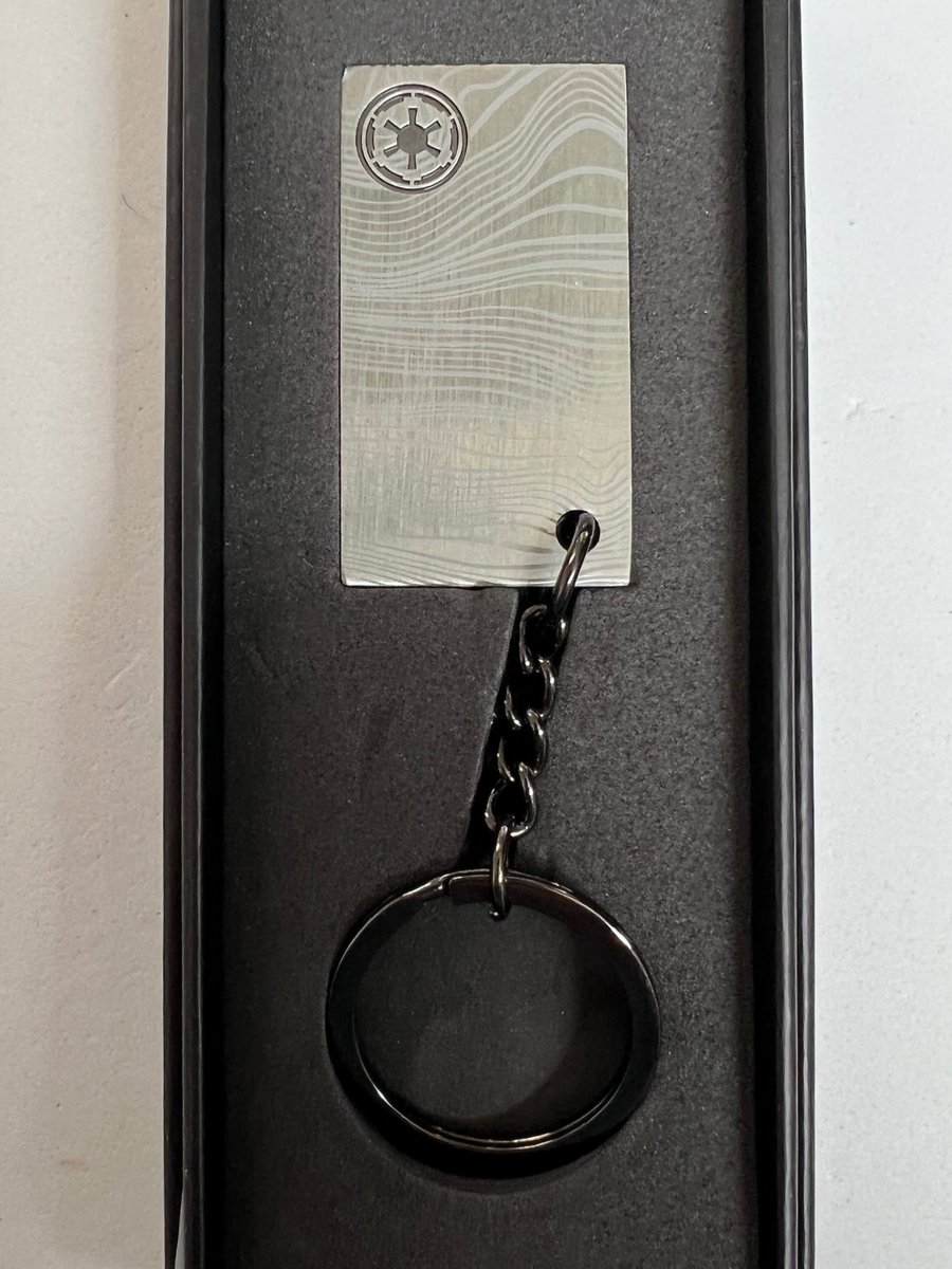 A LEGO VIP keychain, shaped like a silver grey bar of beskar steel from The Mandalorian. It has an Imperial insignia stamped on it in the upper left hand corner. 