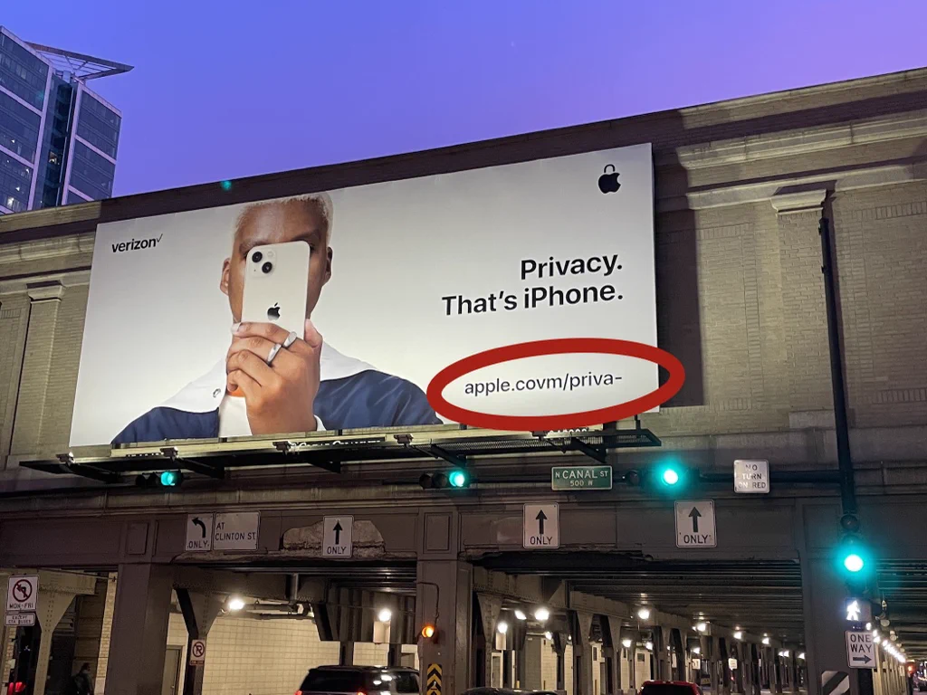 Billboard advertisement showing a person with dark skin and short hair holding a white iPhone in front of their face. In the top left is the Verizon logo, and the top right is the Apple logo modified to look like a padlock. The large text says "Privacy. That's iPhone." The bottom right corner says "apple.covm/priva-"; a red circle has been drawn on the image surrounding this URL