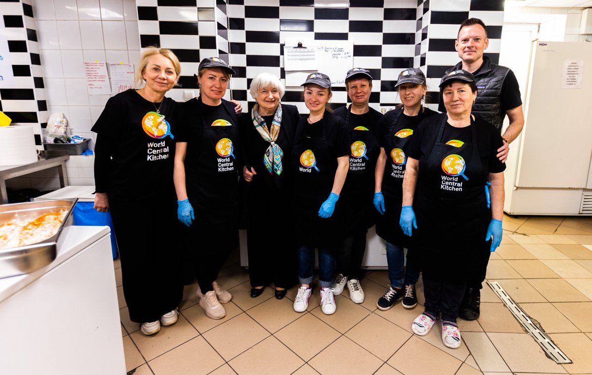 Secretary Yellen and Nate Mook, World Central Kitchen CEO, stand with six refugee volunteers in the kitchen of the Warsaw meal site of World Central Kitchen. The volunteers are wearing WCK aprons and caps.