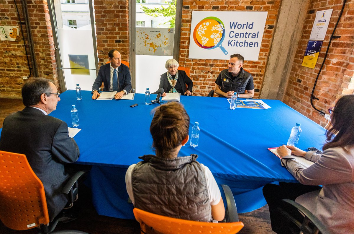 Secretary Yellen sits at a rectangular table with Nate Mook (WCK CEO), Matteo Patrone (European Bank for Reconstruction and Development), Gallina Vincelette (World Bank Group), and other leaders from World Central Kitchen's Warsaw location. A WCK sign is visible in the background.