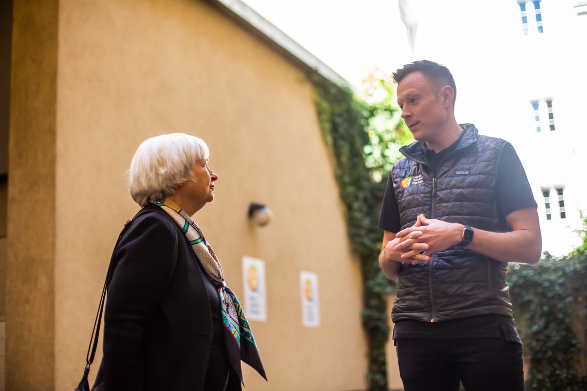 Secretary Yellen speaks with Nate Mook, CEO of the World Central Kitchen. They are facing each other and in conversation. They are outside, the side facade of a building and some plants are visible behind them.