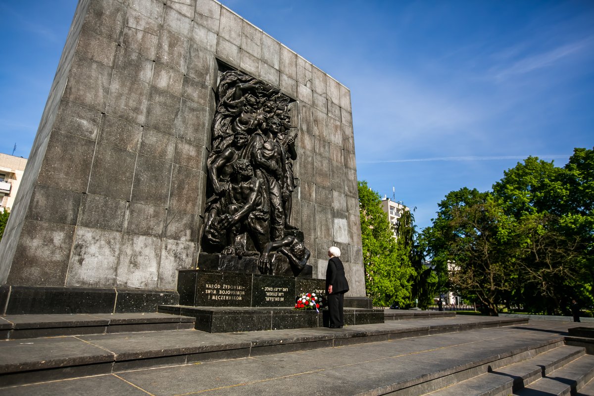 Secretary Yellen places flowers at the base of the Monument to the Ghetto Heroes, which commemorates the 1943 Warsaw Ghetto Uprising. The monument is a large square stone structure with sculpture in the middle depicting a tableau of seven figures. Trees and the sky can be seen in the background.