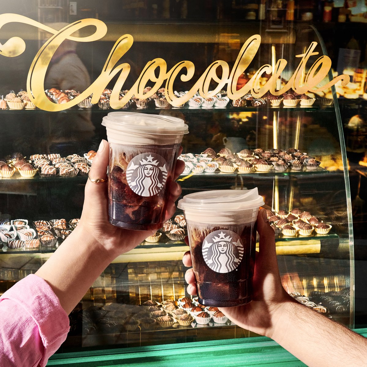 Two hands each hold out their own Chocolate Cream Cold Brew in front of a classic candy shop’s window display full of chocolates.