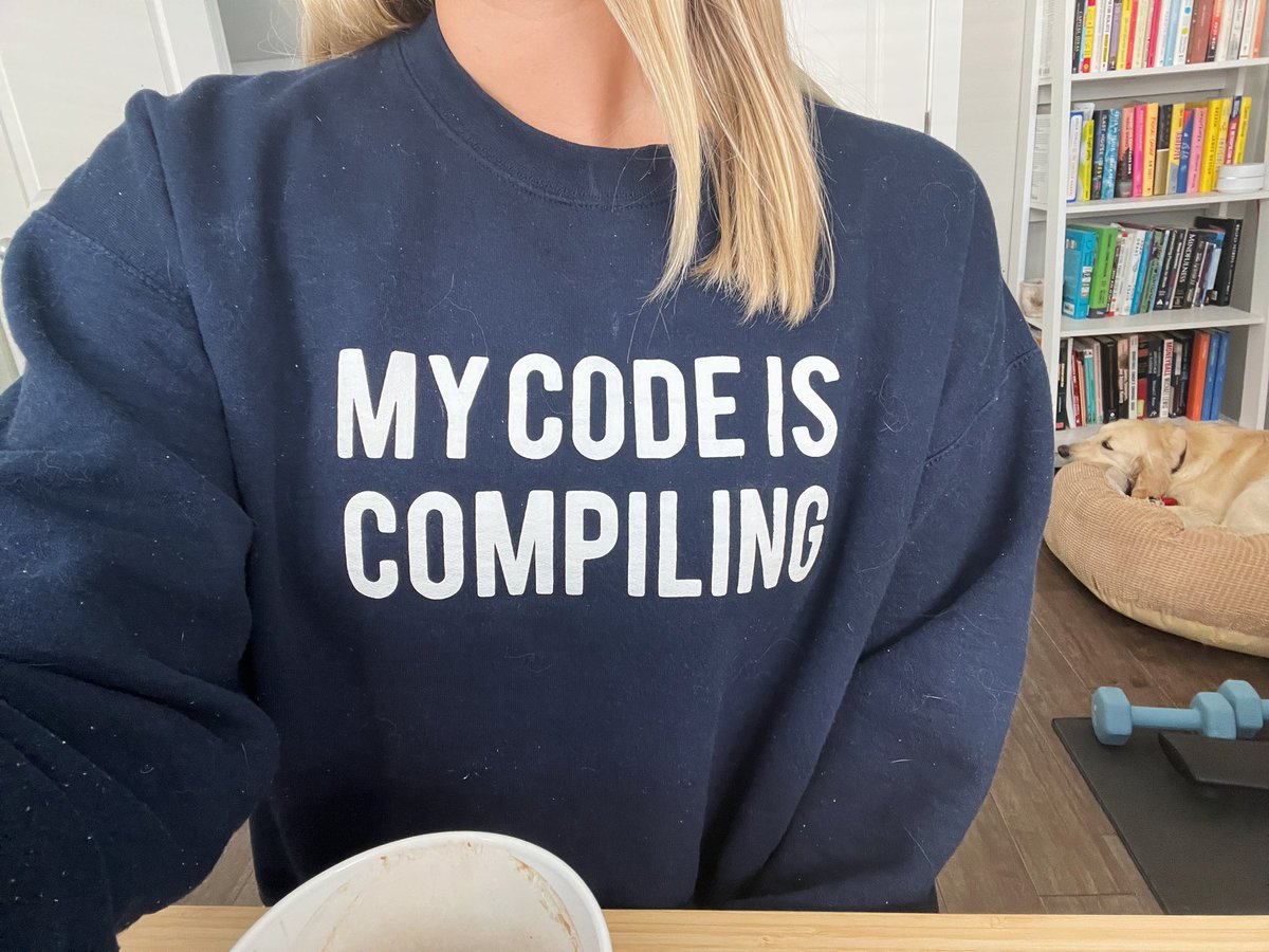 “My code is compiling” on a dark blue sweatshirt covered in cat hair with a bookshelf, a cute golden retriever in the background, and blue weights rarely used.