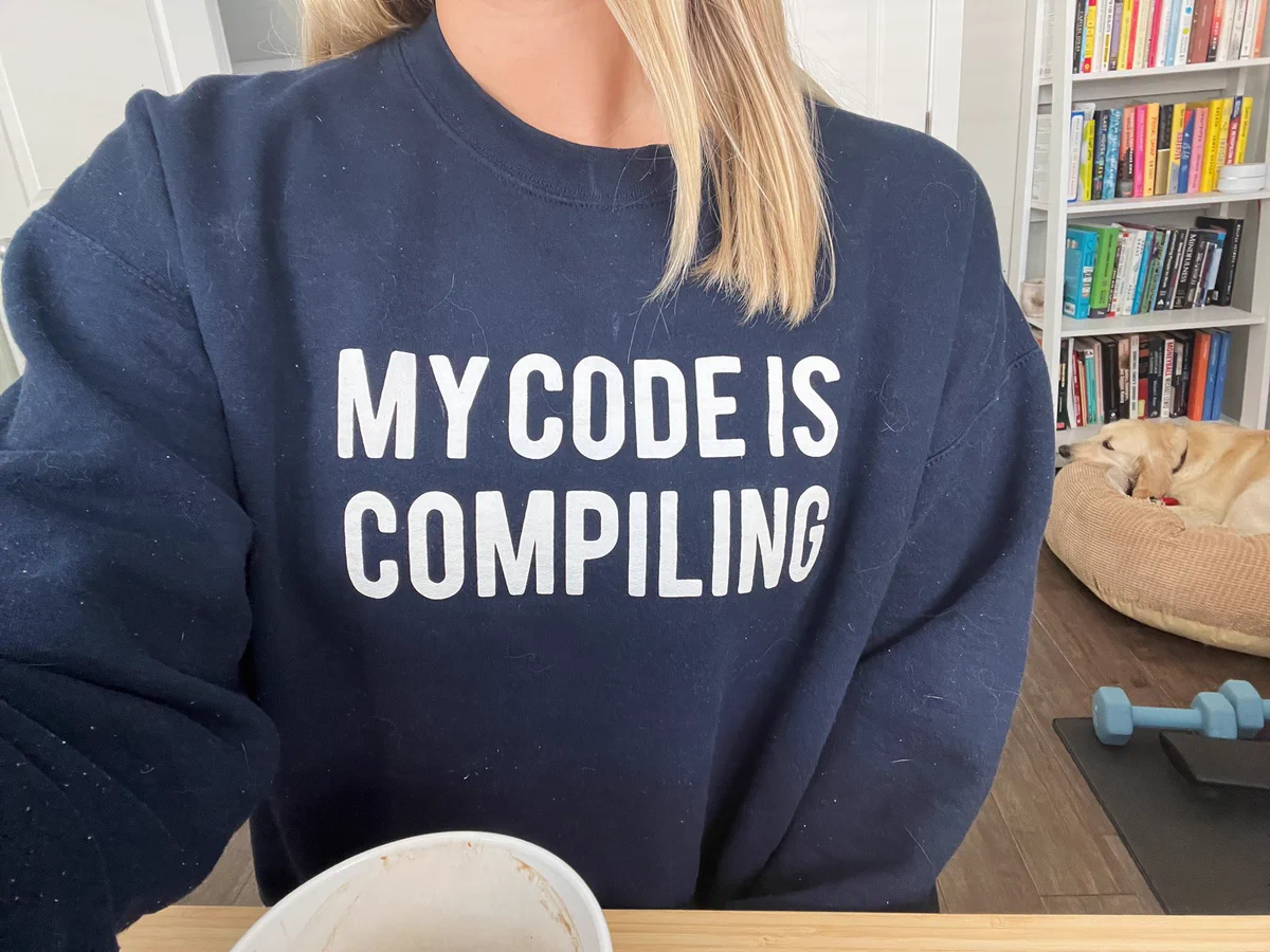 “My code is compiling” on a dark blue sweatshirt covered in cat hair with a bookshelf, a cute golden retriever in the background, and blue weights rarely used.