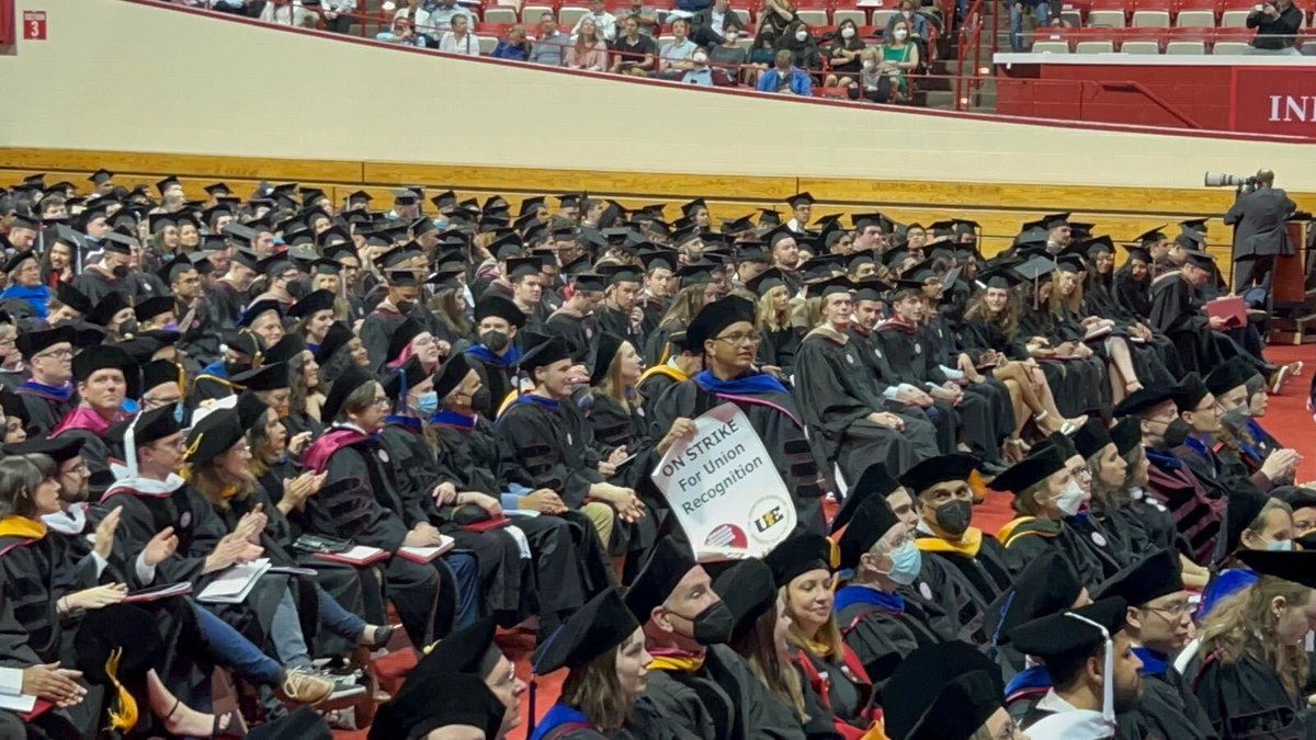 Crowd of graduating PhDs with one person standing up and holding a picket sign that reads: ON STRIKE for union recognition