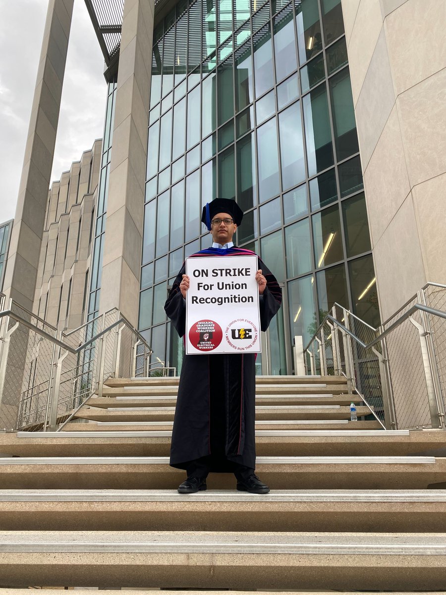Person in a doctoral commencement robe stands in front of the Luddy Building holding a picket sign that reads: ON STRIKE for union recognition