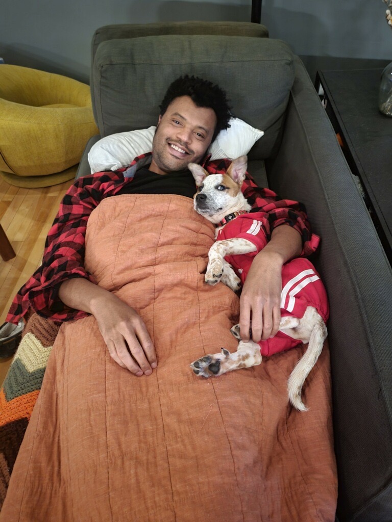 A small brown-and-white speckled puppy with a brown patch over its ears snuggles into a man with dark hair who is covered in a blanket. The pup is wearing a red and white striped set of pajamas, which accent the man's flannel PJ's quite nicely. The man has a big, beautiful grin on his face as he snuggles his new dog. 