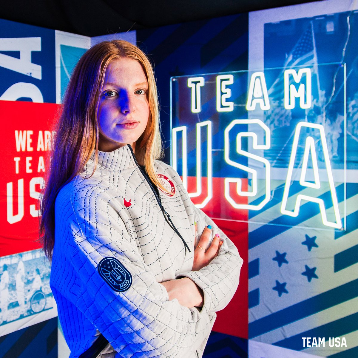 Image of swimmer Lydia Jacoby on a Team USA neon light set wearing a Team USA jacket and crossing her arms 
