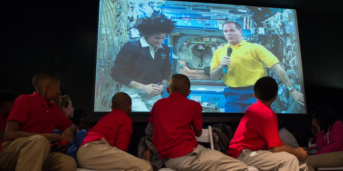 A group of students sit in a row of chairs in an auditorium, and gaze up at a large screen showing two astronauts answering questions during a live, downlink event