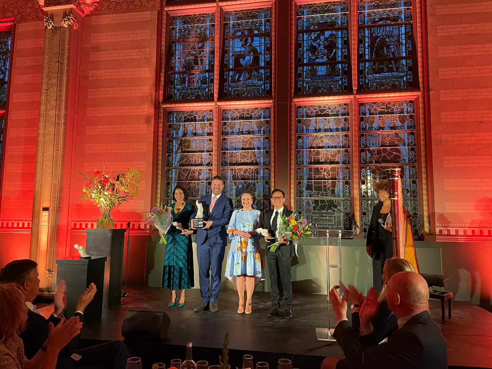 De winnaar en andere genomineerden voor de Young Captain Award staan op het podium met Alexandra van Huffelen. Op de achtergrond een groot glas in lood raam