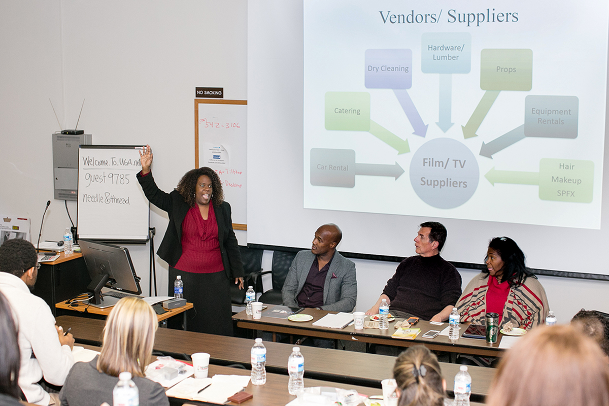 A SBDC staff member addresses attendees of a film panel that are there to learn more about how smal businesses can get involved in Georgia's film industry. The panelist modertor is standing to the left of a table at the front of a classroom. There are three panelists seated at a table in front of a projector screen. Panel attendees are seated facing the panel, with their backs to the camera.