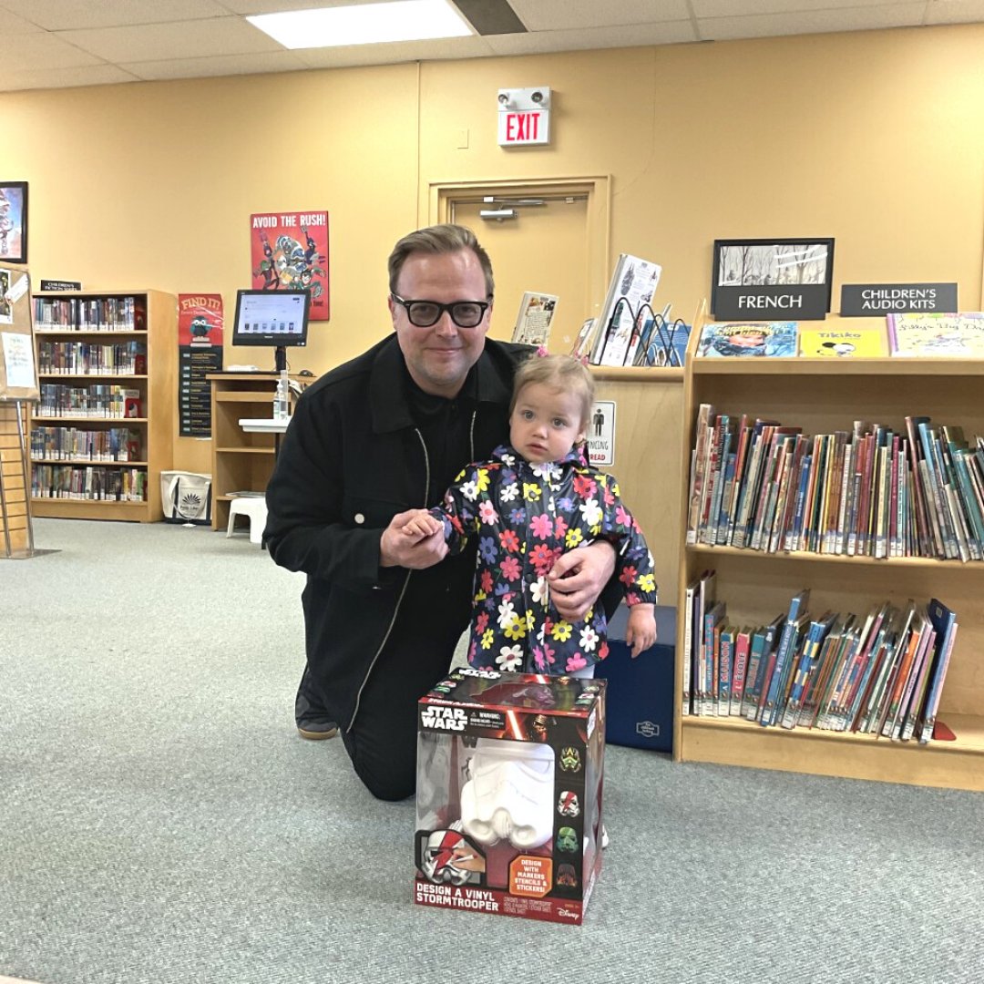 Toddler and father posed in front of a toy bust of a Stormtrooper. In a library setting.