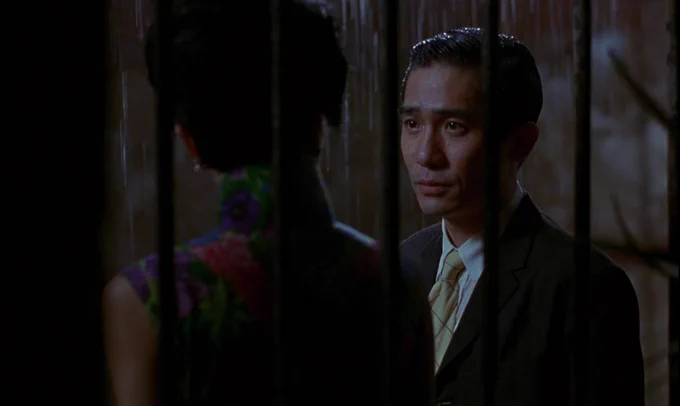 A film still from IN THE MOOD FOR LOVE. A man stands in an alleyway with a woman. He looks at her with a pained look on his face. Rain falls around them.