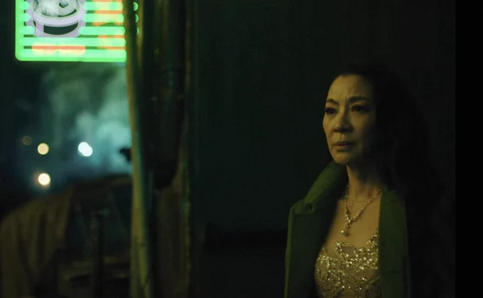 A film still from EVERYTHING EVERYWHERE ALL AT ONCE. A woman wearing an elaborate gown and fur trench stands in an alleyway. She appears uneasy, coloured by green light.