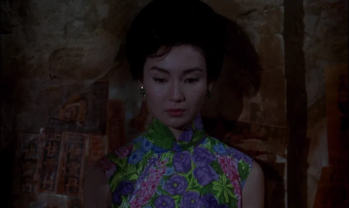 A film still from IN THE MOOD FOR LOVE. A woman rests her back against the wall of an alleyway. Saddened, she casts her view down. 